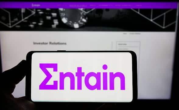 A person displaying the Entain logo on their smartphone against the company's website loaded on a computer screen.