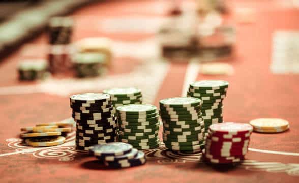 Poker chips stacked at a full poker red-felted table. The chips are in focus whereas the background is slightly blurred.