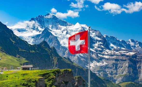 Switzerland national flag in the daylight against a background of a majestic Swiss mountain.