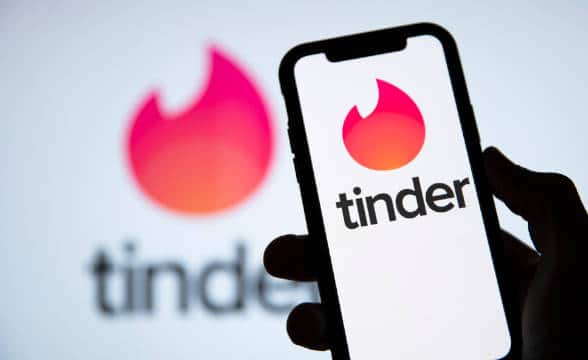 A person holding the Tinder social app, with the app loaded on their smartphone against a background with the company's logo.