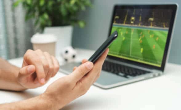 A man holding a smartphone with a laptop playing a soccer game in the background.