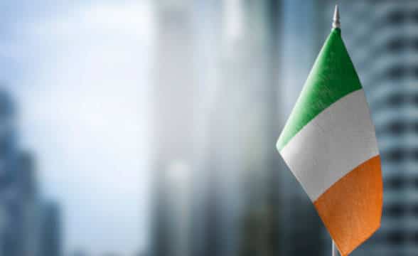 A small flag of Ireland on a pole with blurred urban background