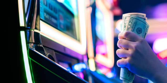 Close up photo of a hand holding money in front of a slot machine, symbolizing jackpot